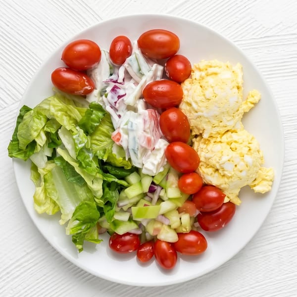 A white plate with sections of lettuce, cherry tomatoes, cucumber salad, and egg salad.