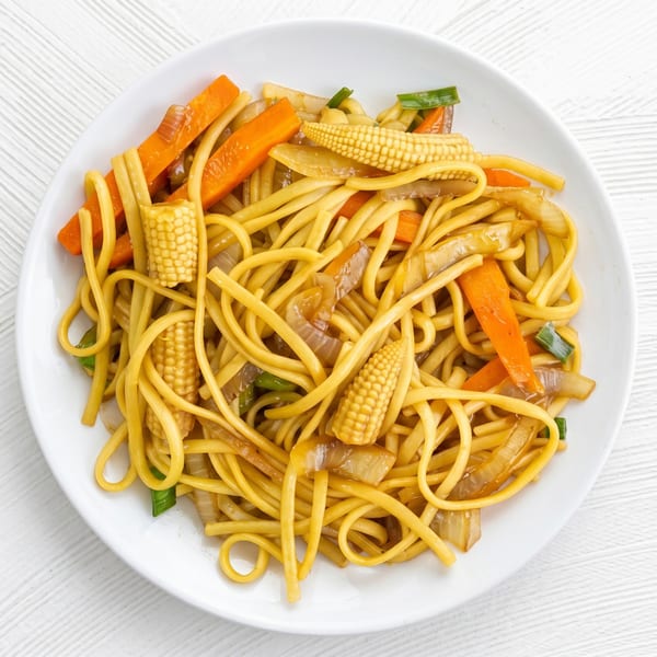 A white plate filled with yellow noodles, carrots, baby corn, and onions in a sauce.